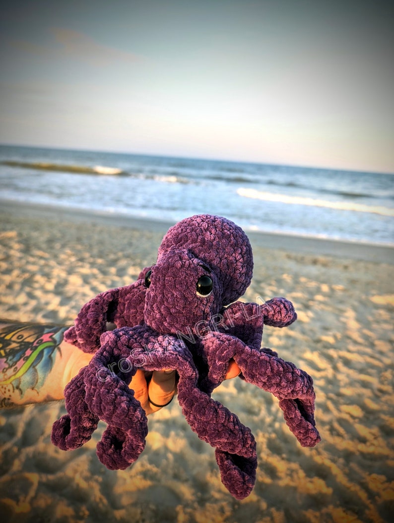 May include: A purple crocheted octopus plush toy with large black eyes. The toy is being held by a hand with a tattoo on the beach.