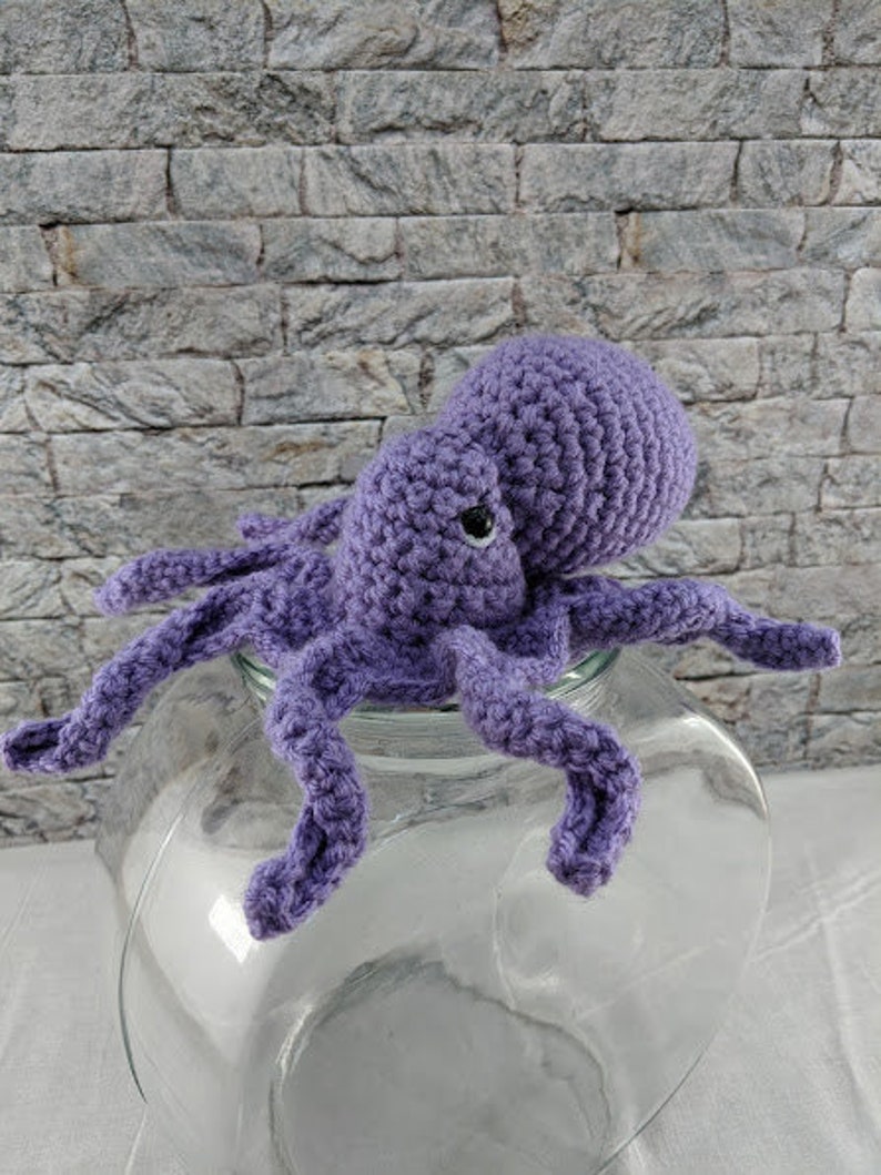 May include: A crocheted purple octopus with eight tentacles, sitting on a clear glass jar.