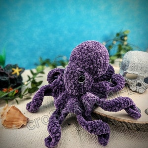 May include: A purple crocheted octopus plush toy with a black eye. The octopus is sitting on a wooden surface with seashells around it.