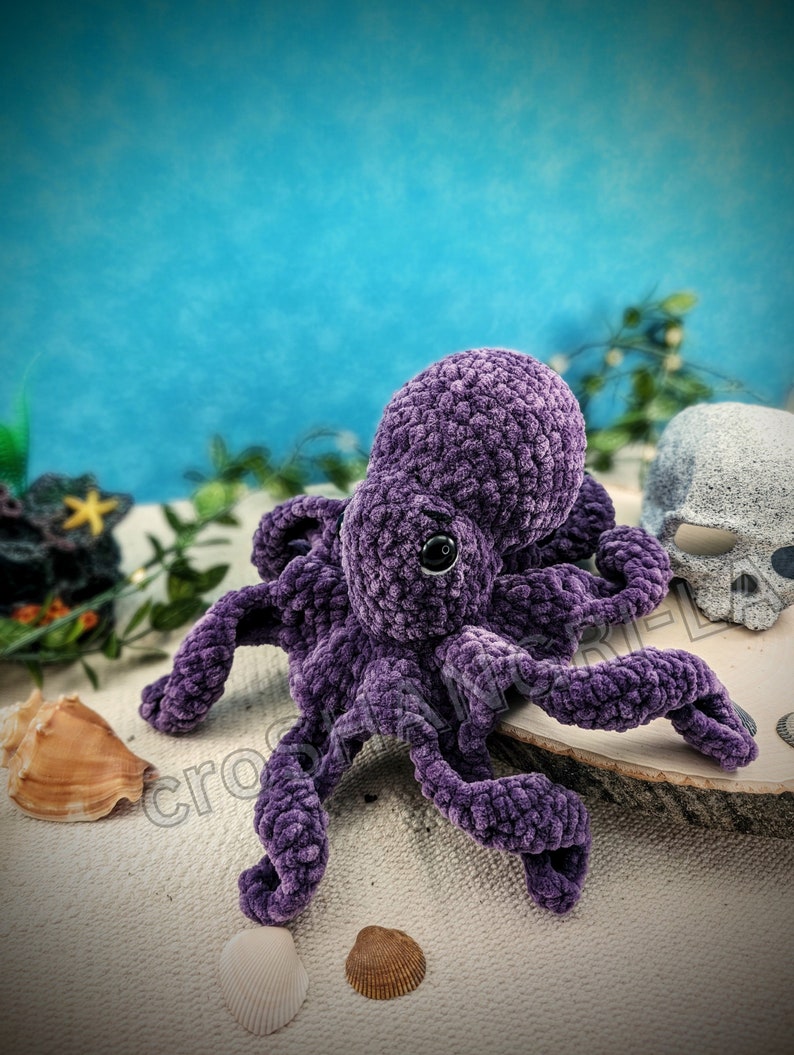 May include: A purple crocheted octopus plush toy with a black eye. The octopus is sitting on a wooden surface with seashells around it.