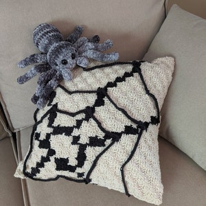 K&ouml;nnte beinhalten: Ein geh&auml;keltes Kissen mit einem schwarzen und wei&szlig;en Spinnennetz-Design. Ein graues Pl&uuml;sch-Spinnenspielzeug sitzt auf dem Kissen.