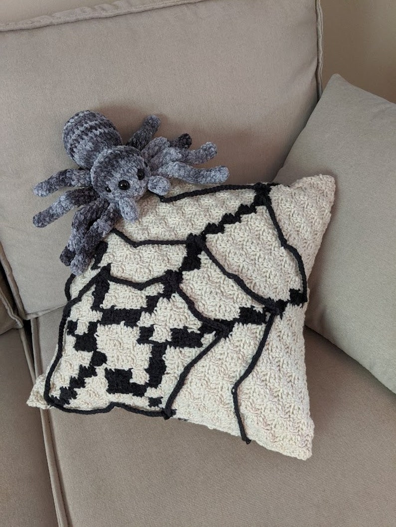K&ouml;nnte beinhalten: Ein geh&auml;keltes Kissen mit einem schwarzen und wei&szlig;en Spinnennetz-Design. Ein graues Pl&uuml;sch-Spinnenspielzeug sitzt auf dem Kissen.