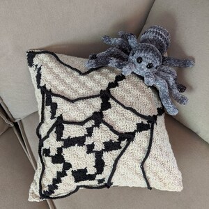 K&ouml;nnte beinhalten: Ein geh&auml;keltes Kissen mit einem schwarzen und wei&szlig;en Spinnennetz-Design. Eine pl&uuml;schige graue Spinne sitzt auf dem Kissen.