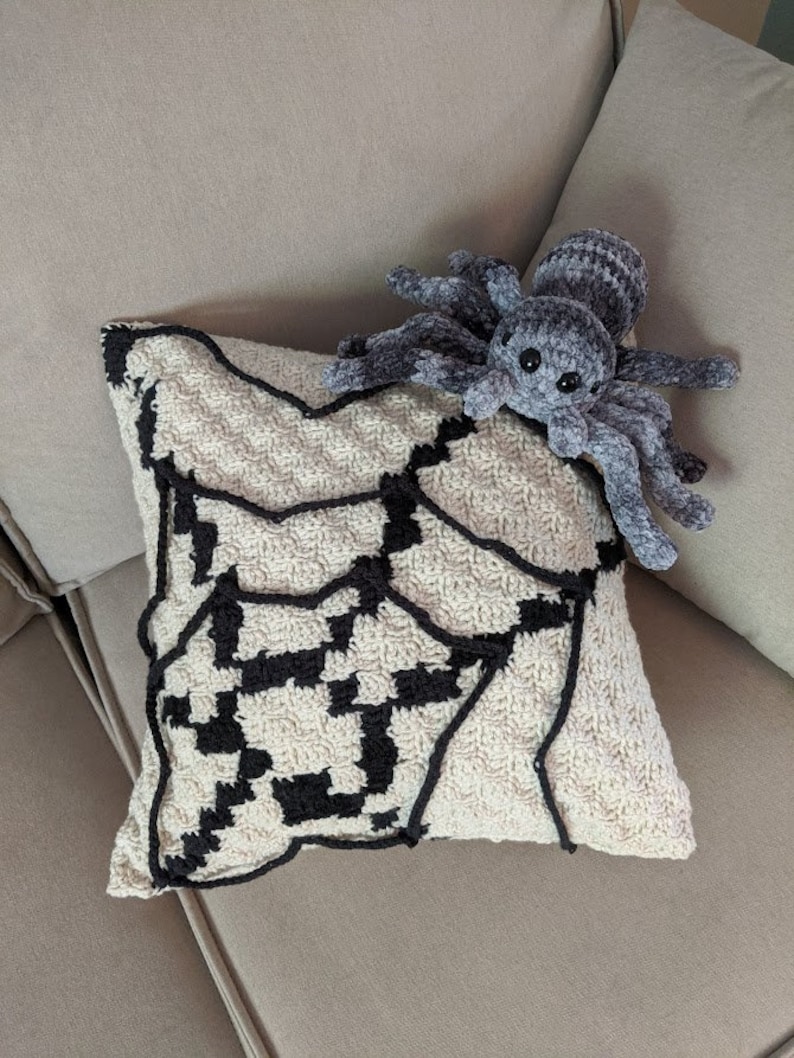 K&ouml;nnte beinhalten: Ein geh&auml;keltes Kissen mit einem schwarzen und wei&szlig;en Spinnennetz-Design. Eine pl&uuml;schige graue Spinne sitzt auf dem Kissen.