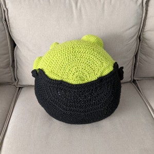 K&ouml;nnte beinhalten: Ein gr&uuml;nes und schwarzes H&auml;kelkissen in Form eines Kopfes. Das Kissen liegt auf einem hellgrauen Sofa.