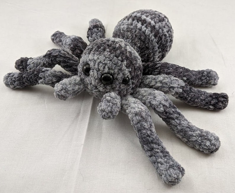 K&ouml;nnte beinhalten: Ein graues und schwarzes geh&auml;keltes Spinnen-Pl&uuml;schtier mit gro&szlig;en schwarzen Augen. Die Spinne hat acht Beine und eine flauschige Textur.