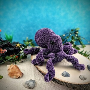 May include: A purple crocheted octopus with black eyes sits on a wooden slice surrounded by seashells.