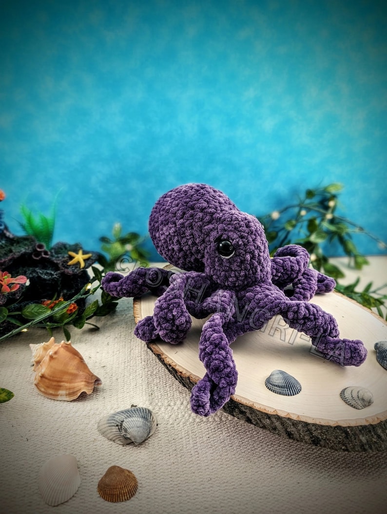 May include: A purple crocheted octopus with black eyes sits on a wooden slice surrounded by seashells.