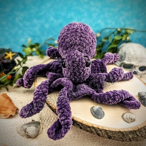 May include: A plush purple crocheted octopus with large black eyes. The octopus is sitting on a wooden slice surrounded by seashells.