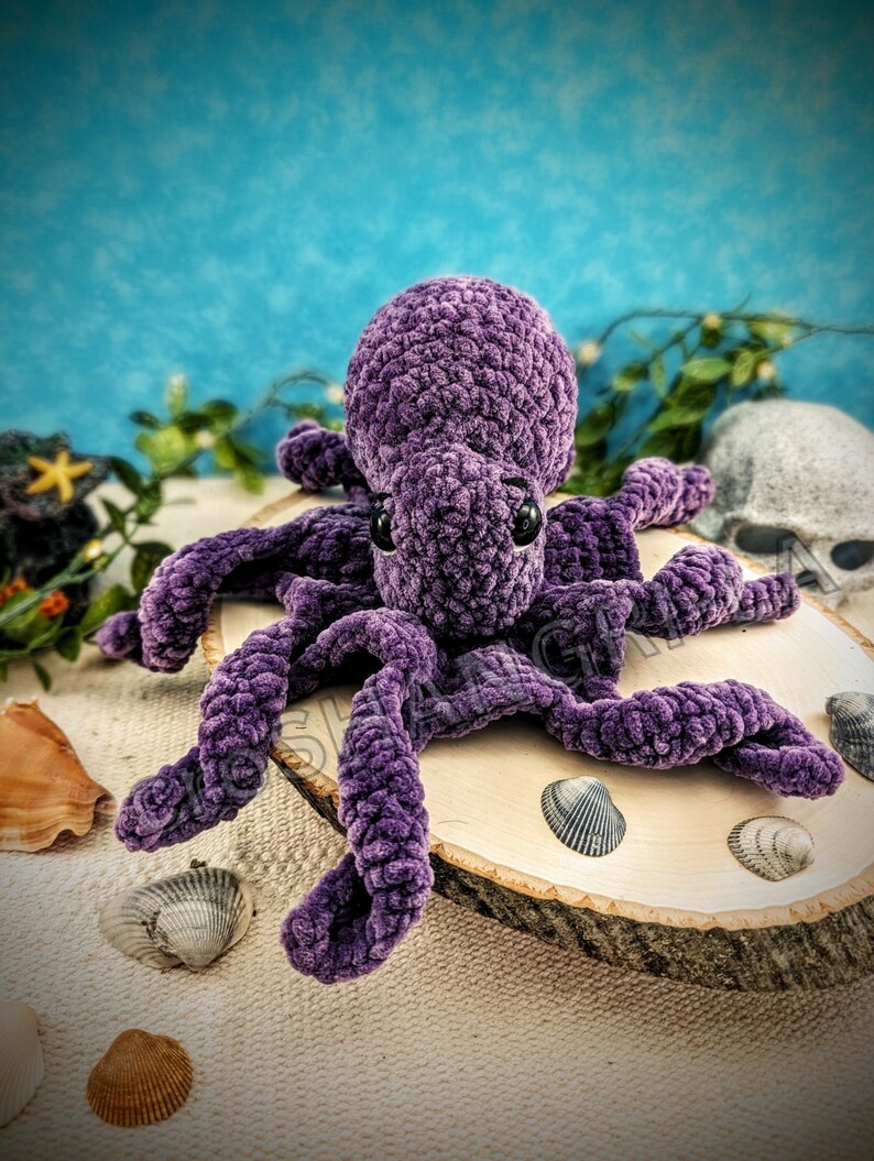 May include: A plush purple crocheted octopus with large black eyes. The octopus is sitting on a wooden slice surrounded by seashells.