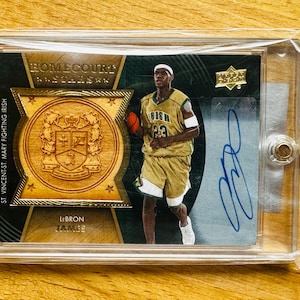Könnte beinhalten: Eine Basketball-Sammelkarte mit LeBron James in einem grün-goldenen Trikot. Die Karte hat einen goldenen Rand und ein Holz-Wappen mit dem Text "Homecourt" und "St. Vincent-St. Mary Fighting Irish". Die Karte ist von LeBron James signiert.