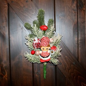 May include: A Christmas decoration featuring a small doll in a red and white outfit, surrounded by faux evergreen branches, red berries, and decorative ornaments. The decoration is mounted on a dark wooden door.