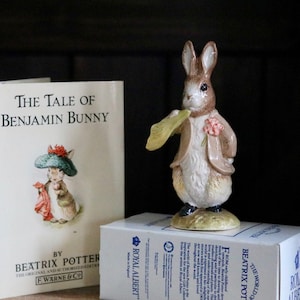 May include: A ceramic figurine of Benjamin Bunny from Beatrix Potter's story, holding a yellow leaf and wearing a brown jacket. Beside it is a copy of "The Tale of Benjamin Bunny" book and a Royal Albert box.