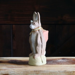 May include: A ceramic rabbit figurine with long ears, painted in shades of brown and tan. The rabbit wears a decorative collar with a light blue and orange ribbon. The figurine is on a light green base, and the background is a dark wooden door.