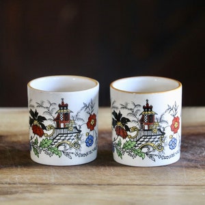 May include: Two white ceramic tumblers with a gold rim and a colorful Asian-inspired design. The design includes a pagoda, flowers, and foliage in red, blue, and green. The tumblers are on a wooden surface.