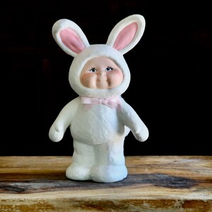 May include: A white ceramic figurine of a child in a bunny costume. The bunny suit has a hood with pink-lined ears and a pink bow tie. The figurine has a painted face with freckles and is standing on a wooden surface.