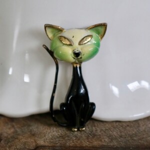 May include: A vintage cat brooch with a black body and a green and cream-colored head. The cat has gold-colored eyes and accents. The brooch is likely made of metal and is a decorative accessory.