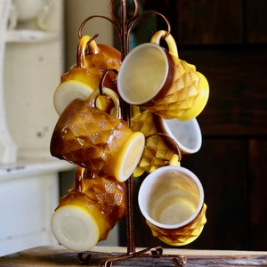 May include: A set of eight amber-colored glass mugs with a textured diamond pattern, hanging on a bronze-colored metal mug tree. The mugs have a yellow exterior and white interior, creating a vintage aesthetic.