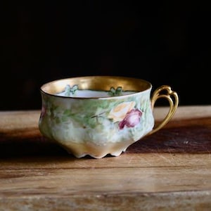 May include: A vintage teacup with a gold rim and handle, featuring a floral design in shades of pink, yellow, and green. The cup sits on a wooden surface, with a dark background. The teacup is a delicate piece of tableware.