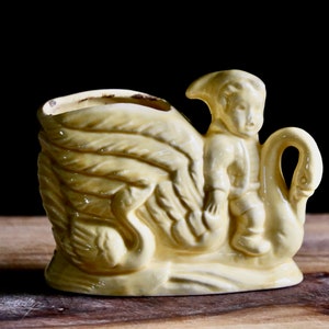 May include: A vintage yellow ceramic planter featuring a swan and a small figure. The planter has a rectangular shape with a detailed swan design on one side and a small figure riding the swan. The planter is approximately 6 inches long.