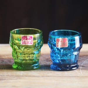 May include: Two small, colorful Viking glass shot glasses. One is green, the other is blue. Both have a skull design and a Viking label. They are sitting on a wooden surface.