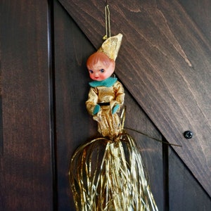 May include: A vintage Christmas ornament featuring a small elf figure with a gold outfit, a green collar, and a red face. The elf has a gold hat and is attached to a gold tassel. The ornament hangs against a dark wood background.