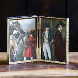May include: A double-sided folding picture frame with a gold-toned metal border featuring a textured pattern. The left panel displays a painting of a person in a red coat with horses, while the right panel shows a painting of a couple in formal attire.