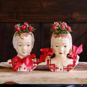 May include: Two vintage ceramic head vases, each featuring a child's face with red bows and a floral arrangement on top. The vases have a plaid pattern on the chest area and are displayed on a wooden surface.