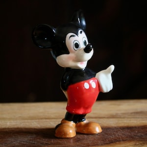 May include: A vintage Mickey Mouse figurine. The figure is depicted in a classic pose, with a black body, red shorts with white buttons, and tan shoes. The figurine has a white gloved hand raised, and a smiling face.