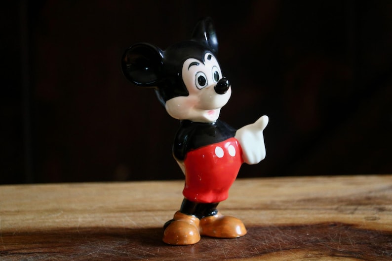 May include: A vintage Mickey Mouse figurine. The figure is depicted in a classic pose, with a black body, red shorts with white buttons, and tan shoes. The figurine has a white gloved hand raised, and a smiling face.