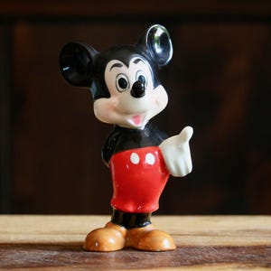 May include: A ceramic Mickey Mouse figurine. The figure has a black body, red shorts with white buttons, and tan shoes. Mickey's face is white with black eyes and ears. He is giving a thumbs-up.