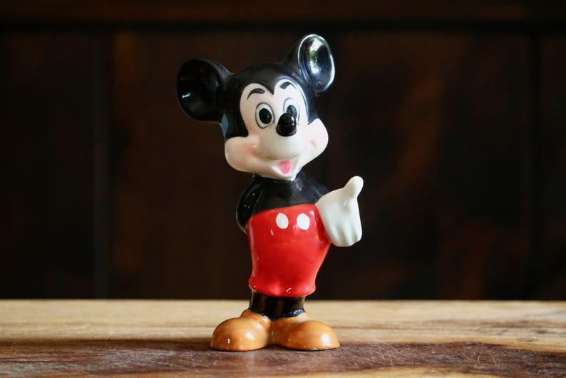 May include: A ceramic Mickey Mouse figurine. The figure has a black body, red shorts with white buttons, and tan shoes. Mickey's face is white with black eyes and ears. He is giving a thumbs-up.