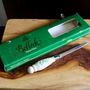 May include: A cheese knife with a white handle featuring a floral design, next to a green box with the text "Belleek, Parian China". The box is open, revealing a white interior. The knife and box rest on a wooden cutting board.