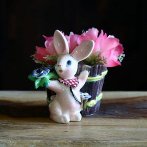 May include: A ceramic bunny figurine with a red and white checkered scarf, holding a bouquet of blue and green flowers. The bunny is in front of a small wooden planter filled with pink flowers. A charming Easter decoration.