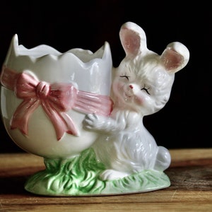May include: A ceramic Easter bunny figurine hugging a cracked egg. The egg is white with a pink bow, and the bunny is light gray with pink accents. The base is green, resembling grass. A charming, decorative Easter-themed item.