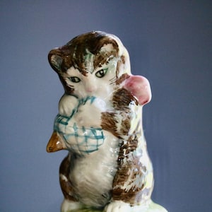 May include: A ceramic figurine of a kitten, primarily white with brown and tan markings. The kitten is holding a blue and white checkered basket and a pink object. The figurine has green eyes and is on a green base.