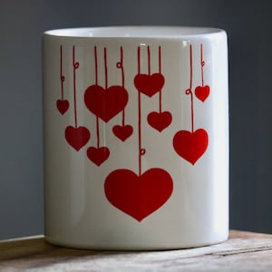 May include: A white ceramic cylinder with a design of red hearts hanging from thin red lines. The hearts vary in size, with the largest at the bottom. The background is a blurred gray.