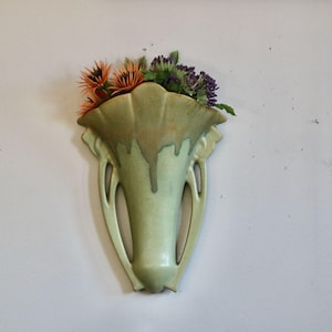 May include: A light green ceramic wall vase with a fluted top and two handles, filled with orange and purple flowers. The vase has a drip glaze effect and is mounted on a white wall.