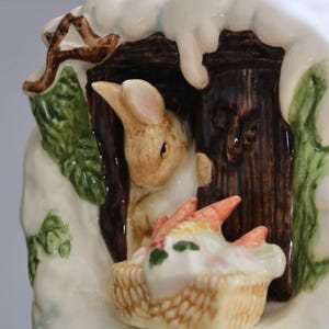 May include: A ceramic figurine featuring a bunny peeking out of a wooden structure, with a basket of carrots in front. The figurine is painted with white, brown, green, and orange colors, and has a snowy roof.
