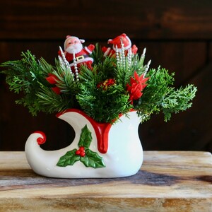 May include: A festive Christmas decoration featuring a white ceramic elf boot filled with greenery, red poinsettias, and two Santa Claus figurines. The boot has a red trim and holly accents, creating a cheerful holiday display.