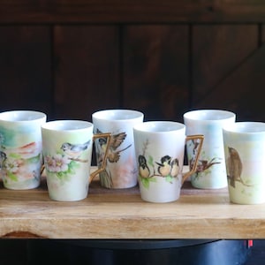 May include: A collection of seven white ceramic mugs with gold-colored handles, each adorned with a unique watercolor painting of birds and floral elements. The mugs are displayed on a wooden tray, showcasing their artistic designs.