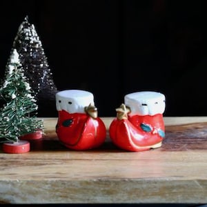 May include: Two red and white ceramic Christmas stocking salt and pepper shakers with gold accents. A small green Christmas tree and red candles are also on the wooden surface. The background is black.