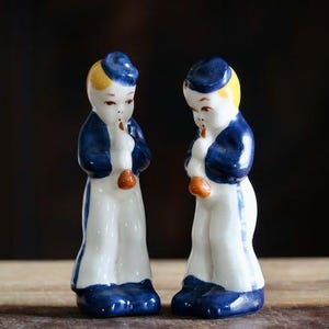 May include: A pair of vintage ceramic figurines depicting figures in sailor-like attire. Each figurine is painted with a blue hat and jacket, white pants, and orange accents. The figures are standing and holding a small object.