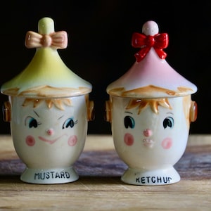 May include: A pair of vintage ceramic condiment jars, one labeled "MUSTARD" and the other "KETCHUP." Each jar is shaped like a smiling face with a conical lid topped with a bow. The mustard jar has a yellow lid and the ketchup jar has a pink lid.