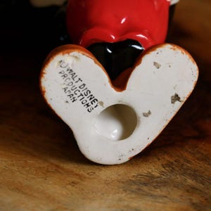 May include: A vintage ceramic figurine of a cartoon character, featuring a red head, black body, and white gloves. The figurine's feet are white with orange accents. The base of the figurine is stamped with "Walt Disney Productions Japan."
