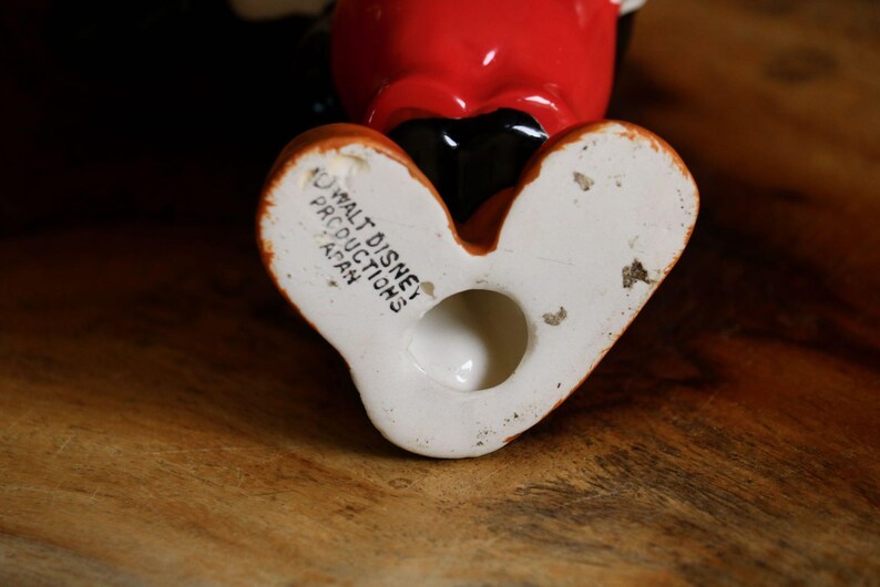 May include: A vintage ceramic figurine of a cartoon character, featuring a red head, black body, and white gloves. The figurine's feet are white with orange accents. The base of the figurine is stamped with "Walt Disney Productions Japan."
