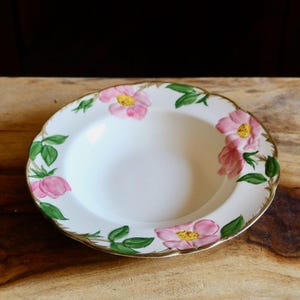 May include: A white ceramic bowl with a scalloped edge, adorned with pink floral and green leaf designs. The flowers have yellow centers and the rim is trimmed with gold. The bowl is set on a wooden surface.