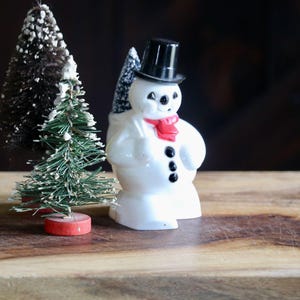 May include: A ceramic snowman figurine with a black top hat, red scarf, and black button details. A small, green Christmas tree with white accents and a red base sits beside the snowman. The items are on a wooden surface.