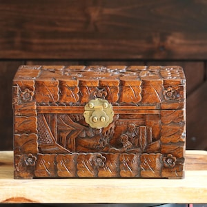 May include: A rectangular dark brown wooden box featuring intricate hand-carved relief scenes of figures and architecture on the front panel. The lid and edges are decorated with carved floral motifs and a central brass latch mechanism.