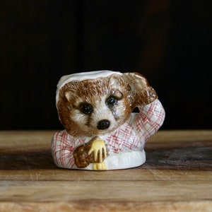May include: A ceramic figurine of a badger wearing a white nightcap and a red and white plaid shirt. The badger is holding a small object in its paw. The figurine has brown and white fur and is set against a dark background.